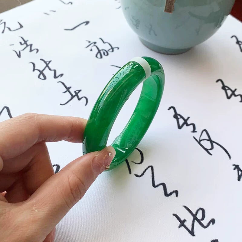Hand holding natural vivid green gradient jadeite bangle over traditional patterned fabric with open book in background 02