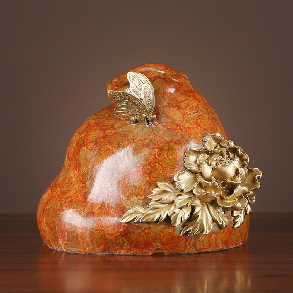 Orange textured decorative sculpture with brass peony flower and butterfly details, displayed on wooden table against dark background