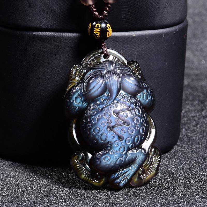 Rainbow obsidian pendant intricately carved into the shape of a toad, adorned with an "Om" bead, suspended from a cord and placed against a dark - colored box backdrop.
