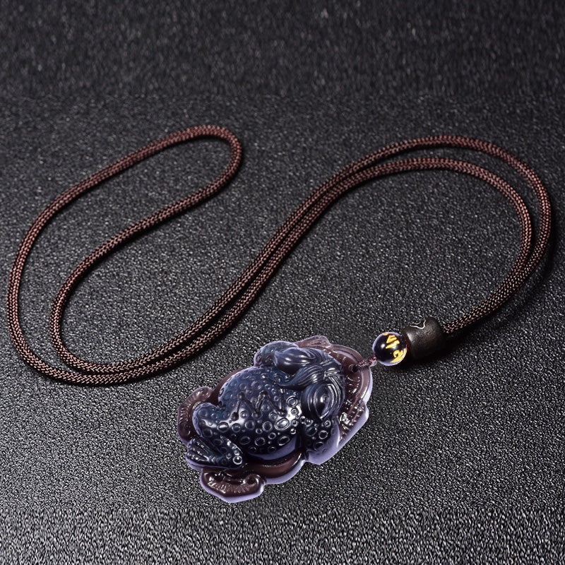 Rainbow obsidian pendant intricately carved into the shape of a toad, adorned with an "Om" bead, suspended from a cord and placed against a dark - colored box backdrop. 01