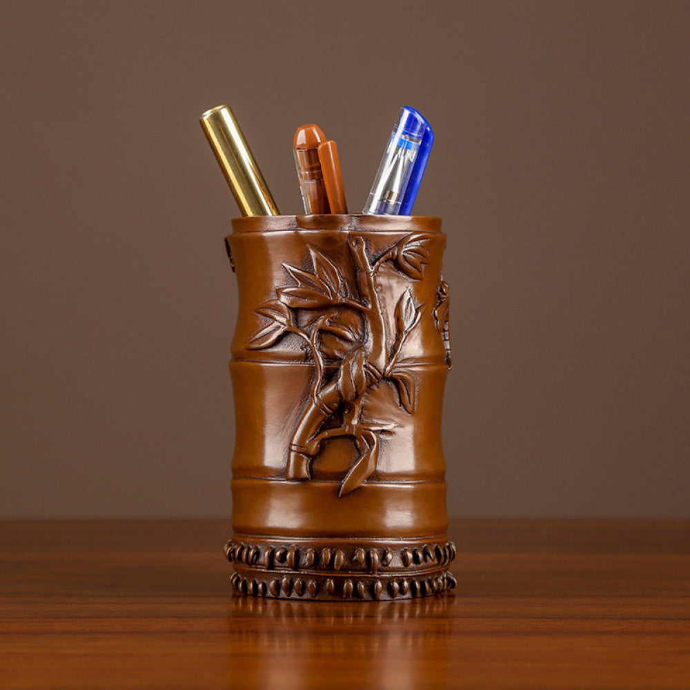Red copper brush pot with bamboo carvings, holding pens, placed on wooden table against brown background