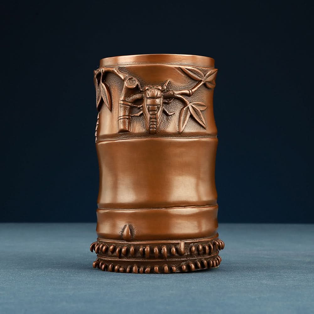 Red copper brush pot with bamboo carvings, holding pens, placed on wooden table against brown background 01