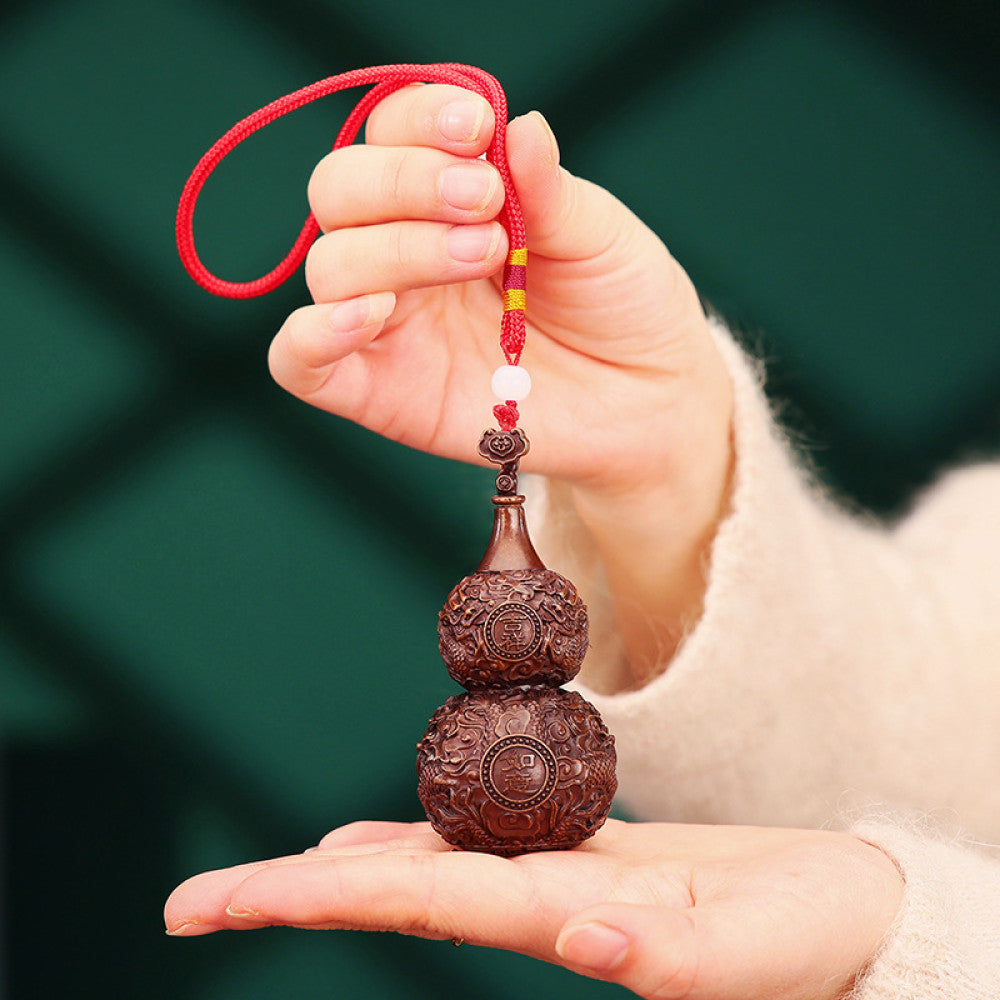 Traditional Chinese carved wooden gourd ornament with intricate patterns and red cord, placed against a blurred green plant background 01