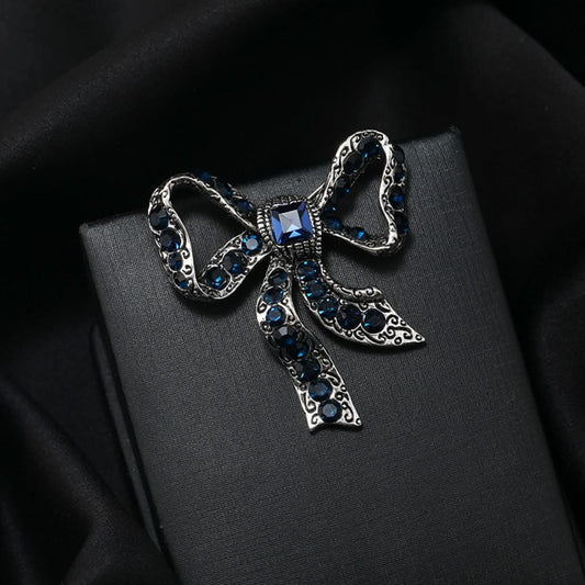 This eye-catching brooch features a classic bow design, crafted with an antiqued gold-tone metal base intricately adorned with vibrant red crystals. A large, faceted red gemstone anchors the center, while delicate filigree detailing adds vintage charm. Perfect for elevating blazers, scarves, or festive outfits, this bold piece brings a touch of glamorous retro style to any look 01