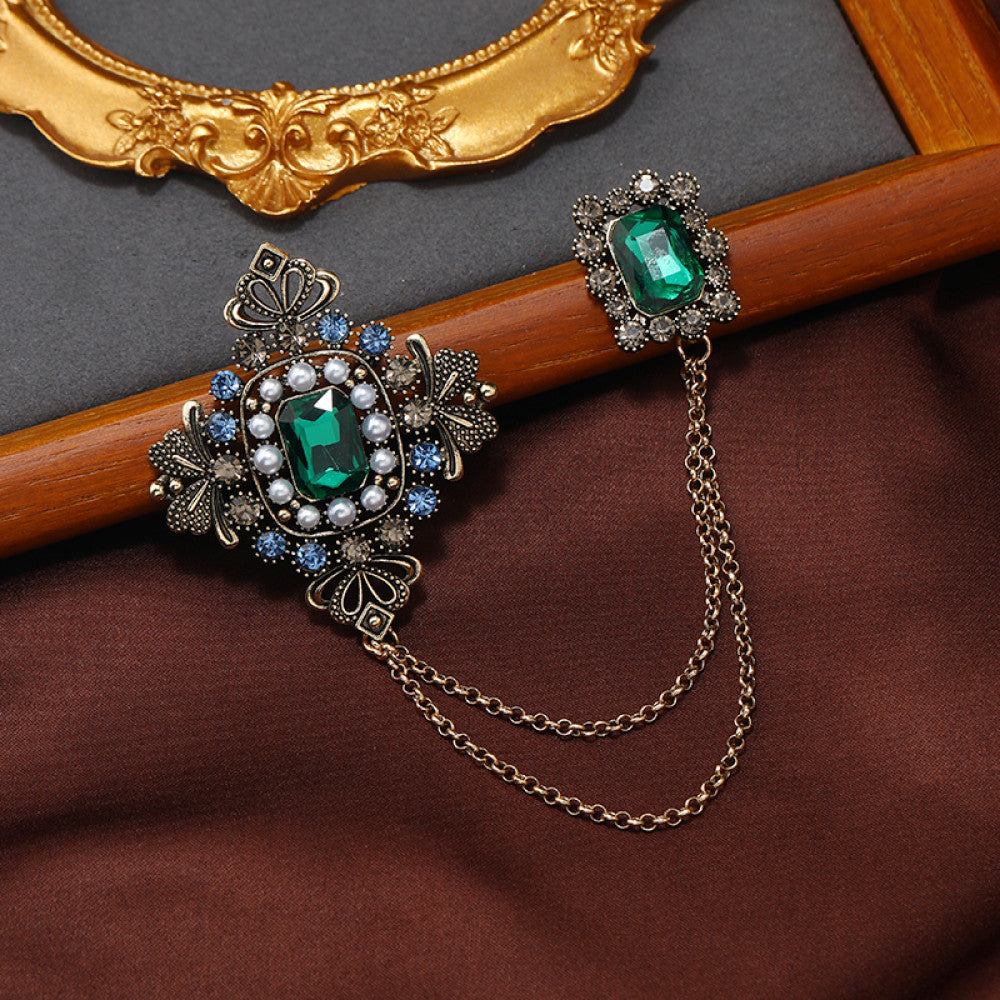 This vintage-style double brooch set features an ornate filigree design with a large emerald-green gem at its center, connected by delicate chains to a smaller matching brooch. Adorned with blue and white accents, it adds sophisticated antique charm to any outfit