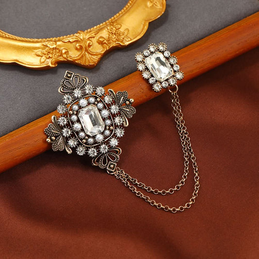 This vintage-style double brooch set features an ornate filigree design with a large emerald-green gem at its center, connected by delicate chains to a smaller matching brooch. Adorned with blue and white accents, it adds sophisticated antique charm to any outfit 01