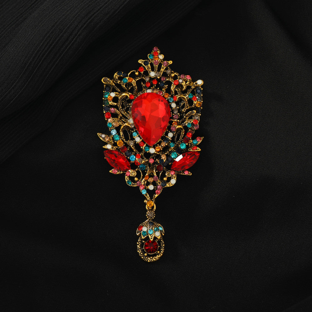 This eye-catching brooch features an ornate, vintage-inspired design with an antiqued gold filigree base. At its center, a large, faceted red crystal takes the spotlight, surrounded by a vibrant array of multicolored crystals and beads. A delicate dangle pendant, adorned with additional gemstones, hangs from the bottom, adding movement and drama. Perfect for elevating blazers, evening gowns, or special occasion outfits, this bold piece exudes opulent, baroque-inspired charm.