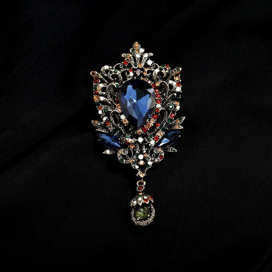 This eye-catching brooch features an ornate, vintage-inspired design with an antiqued gold filigree base. At its center, a large, faceted red crystal takes the spotlight, surrounded by a vibrant array of multicolored crystals and beads. A delicate dangle pendant, adorned with additional gemstones, hangs from the bottom, adding movement and drama. Perfect for elevating blazers, evening gowns, or special occasion outfits, this bold piece exudes opulent, baroque-inspired charm. 01