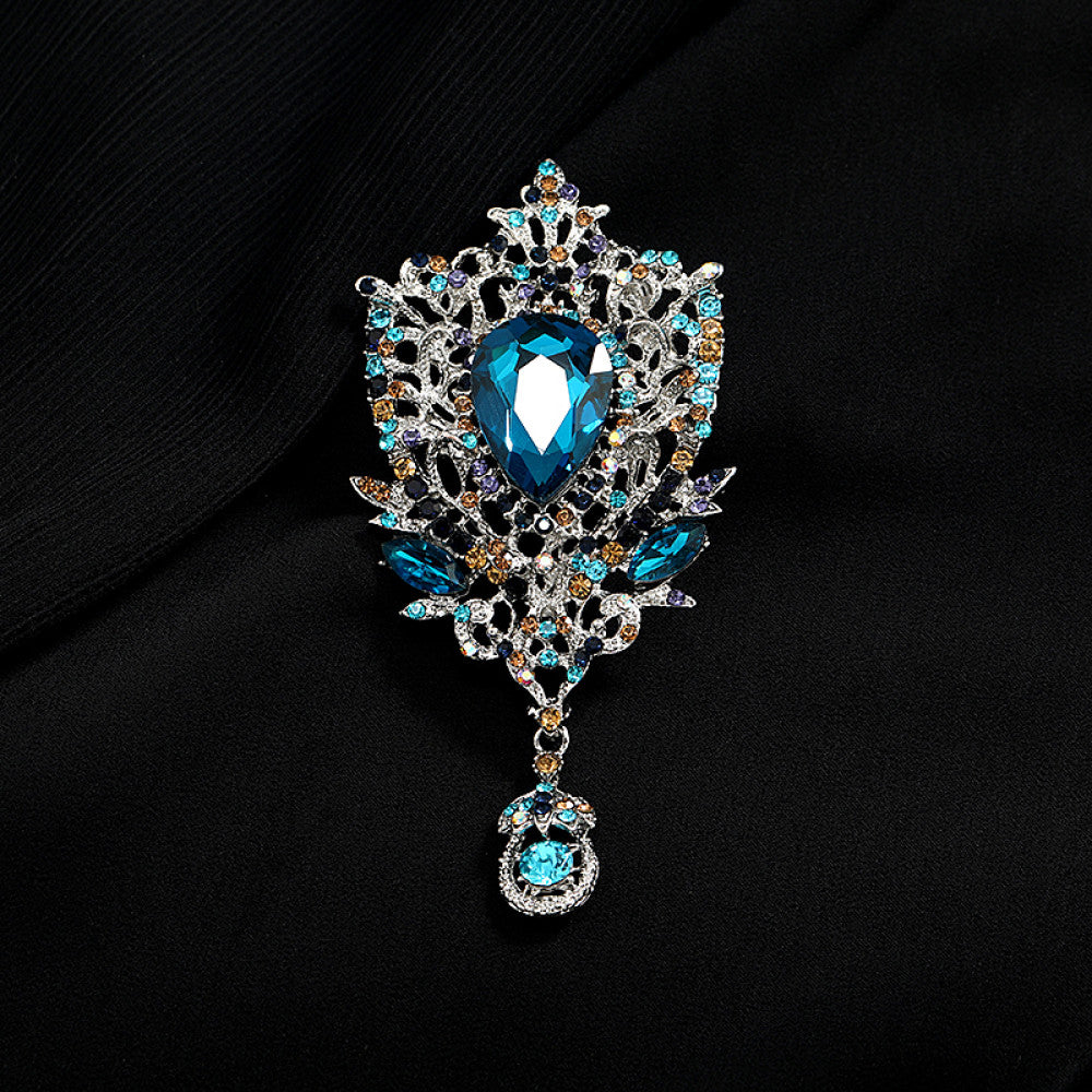 This eye-catching brooch features an ornate, vintage-inspired design with an antiqued gold filigree base. At its center, a large, faceted red crystal takes the spotlight, surrounded by a vibrant array of multicolored crystals and beads. A delicate dangle pendant, adorned with additional gemstones, hangs from the bottom, adding movement and drama. Perfect for elevating blazers, evening gowns, or special occasion outfits, this bold piece exudes opulent, baroque-inspired charm. 02