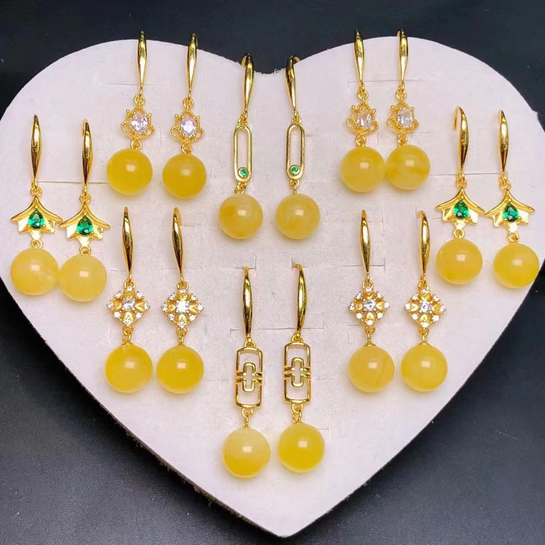 Set of yellow amber earrings with gold - plated hooks and findings, adorned with gemstones like diamonds and emeralds, displayed on a heart - shaped white jewelry holder.