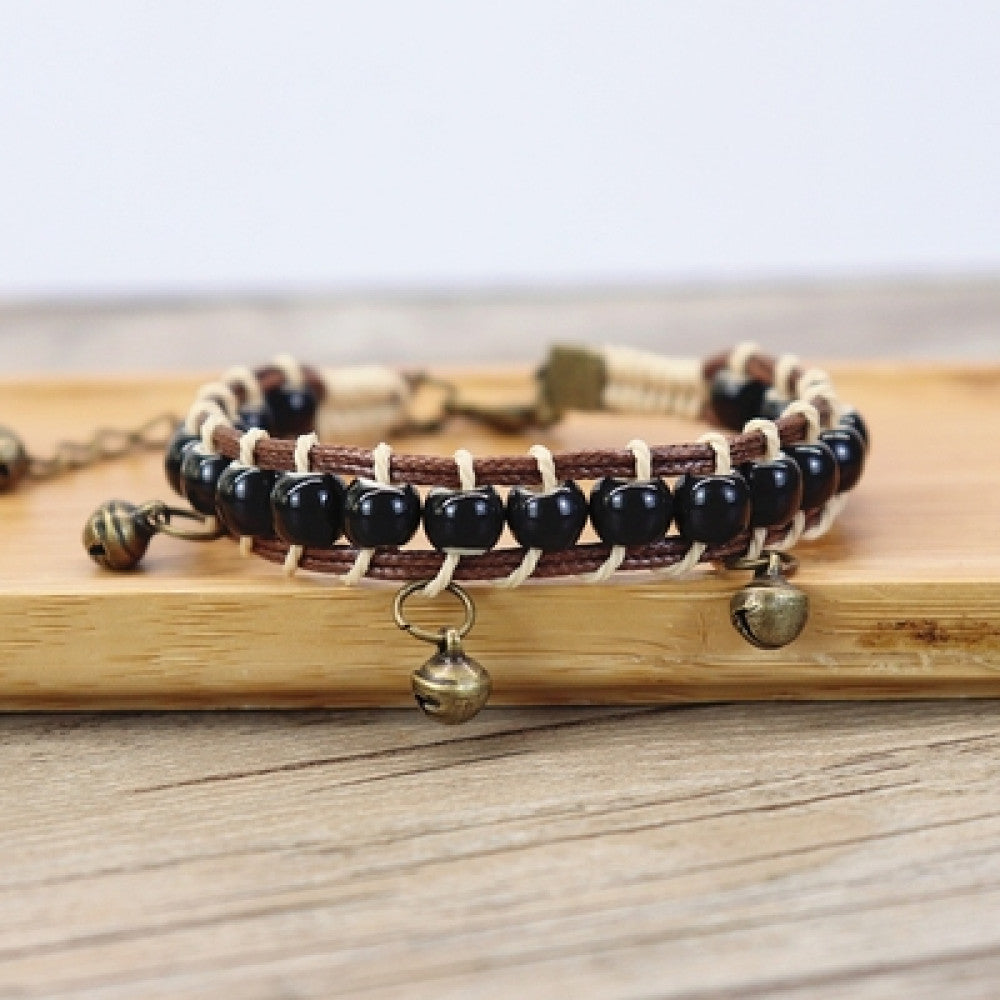 This bohemian bracelet features vibrant multicolored ceramic beads woven onto a brown leather cord, accented with small bronze bell charms. The adjustable design ensures a comfortable fit