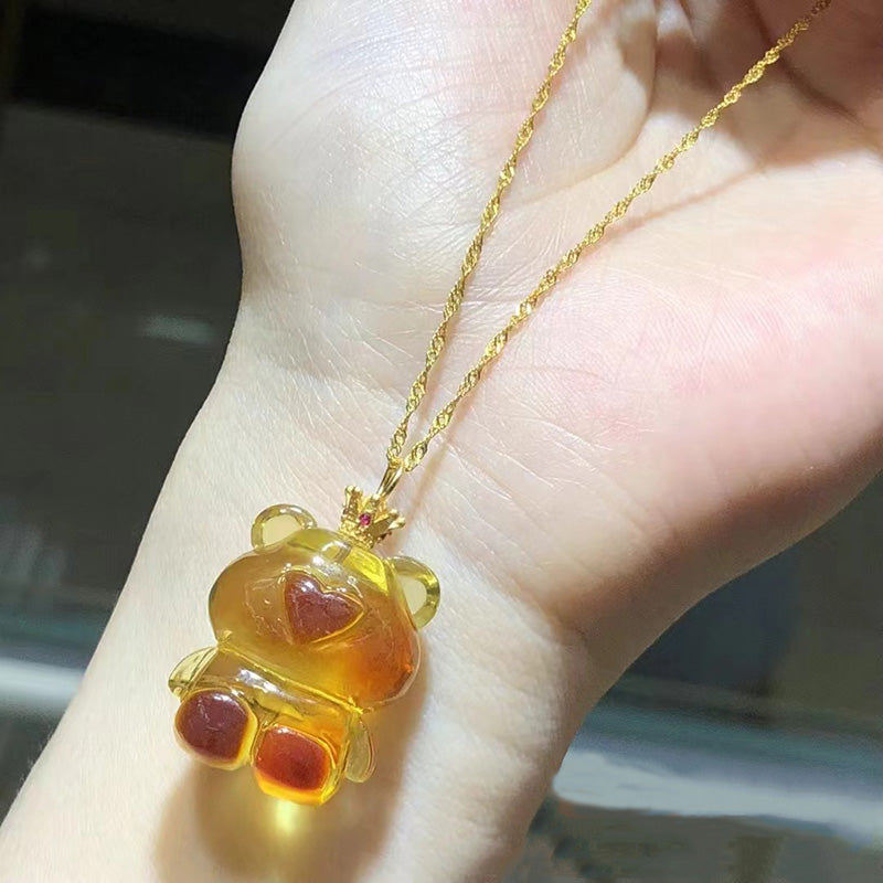 A necklace featuring a carved amber teddy bear pendant with red accents, attached to a gold - toned chain, held in a hand.