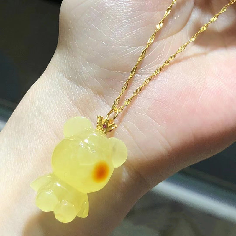 A necklace featuring a carved amber teddy bear pendant with red accents, attached to a gold - toned chain, held in a hand.