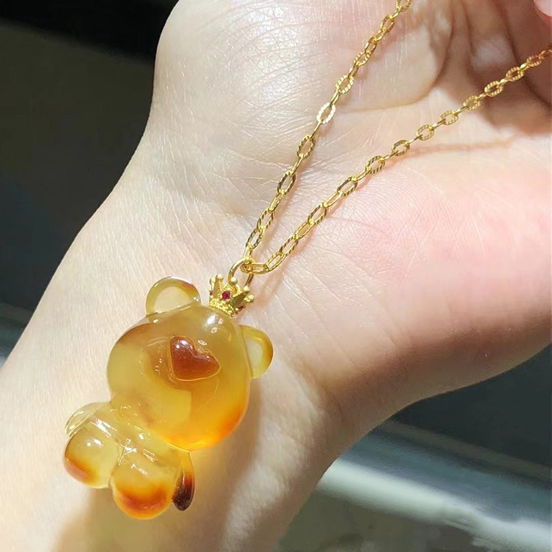 A necklace featuring a carved amber teddy bear pendant with red accents, attached to a gold - toned chain, held in a hand. 02