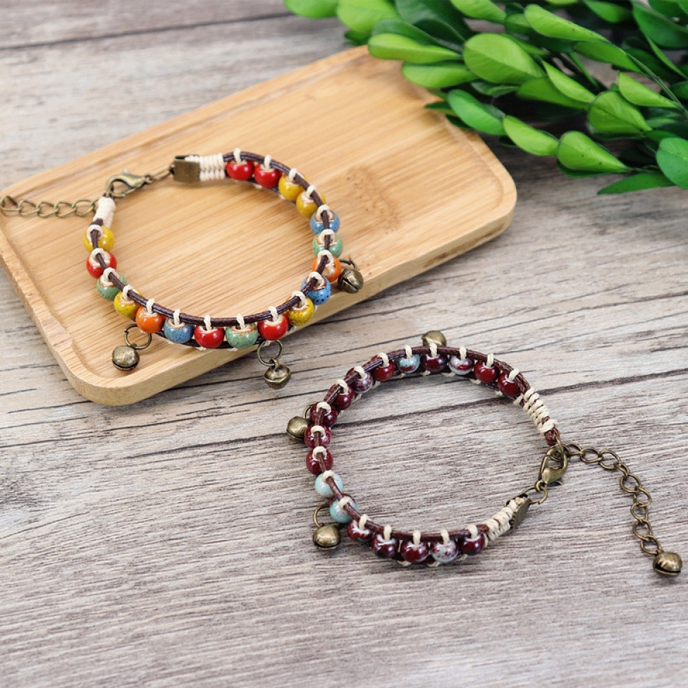 This bohemian bracelet features vibrant multicolored ceramic beads woven onto a brown leather cord, accented with small bronze bell charms. The adjustable design ensures a comfortable fit, adding a playful