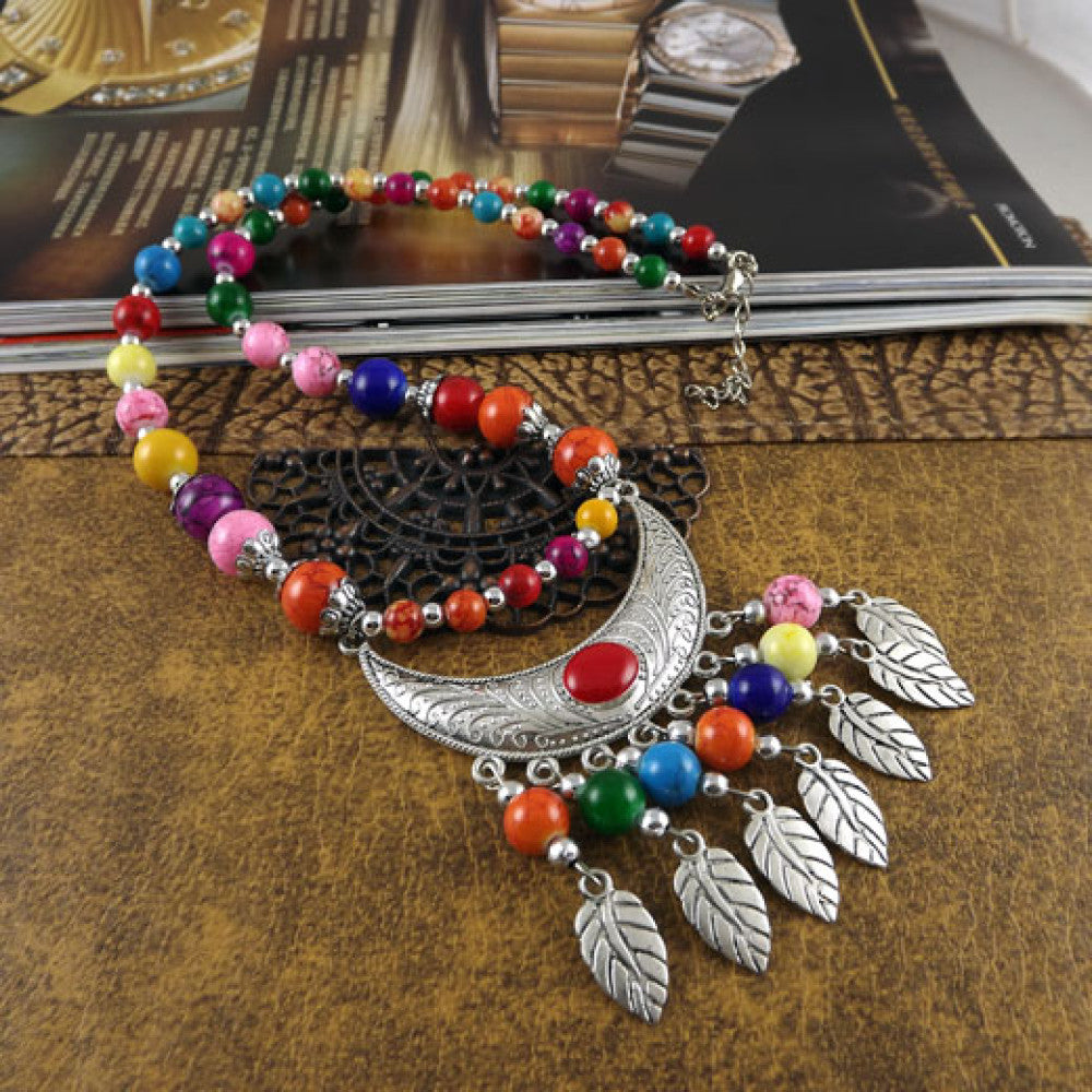 Vibrant colorful beaded necklace with intricate silver-toned leaf tassel and crescent pendant, featuring a mix of bright beads, ideal for bohemian fashion, showcased on a textured surface with floral accents 06
