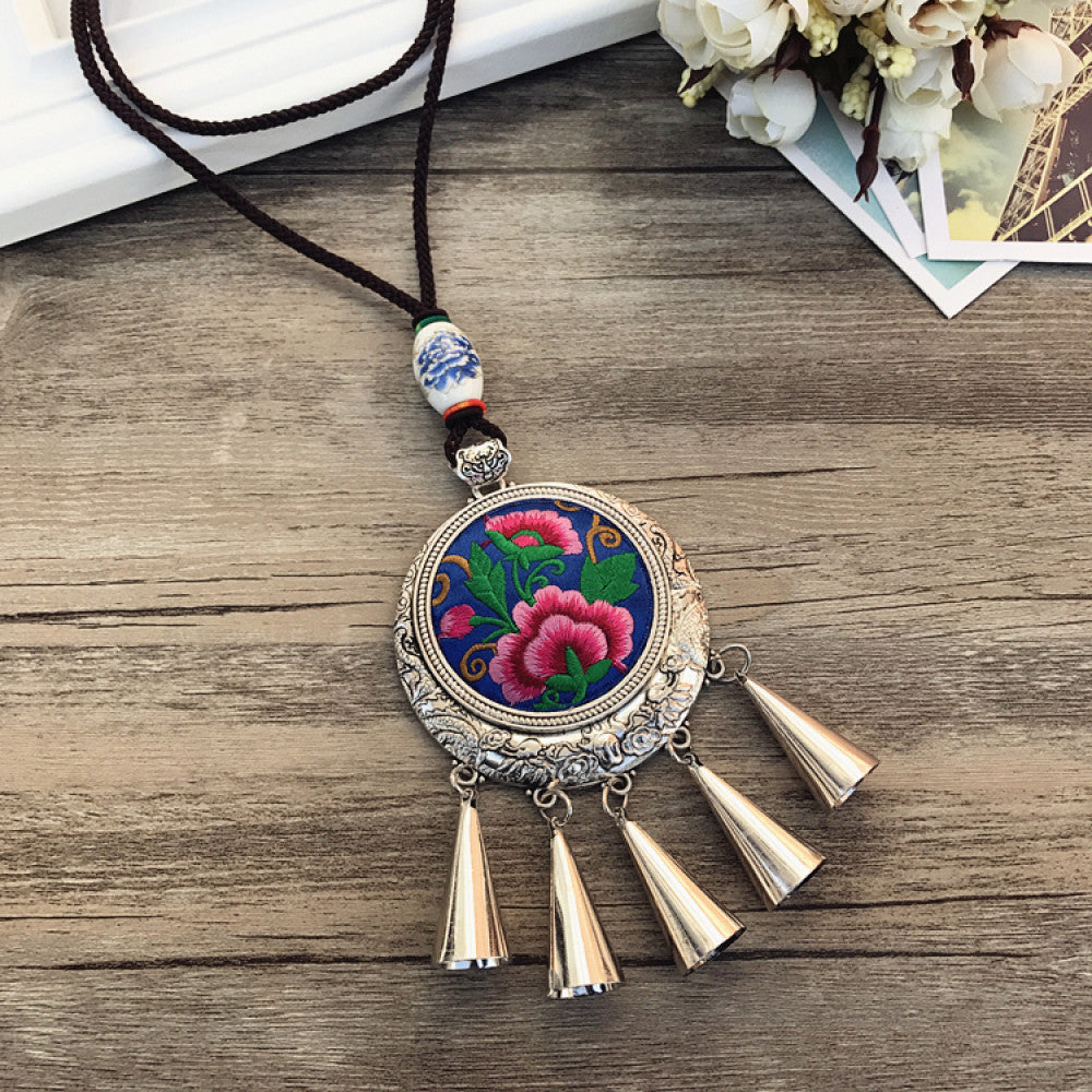 Vintage bohemian ethnic necklace with embroidered floral pendant, silver bells, and ceramic accent, displayed on a wooden surface with flowers, ideal for boho fashion enthusiasts
