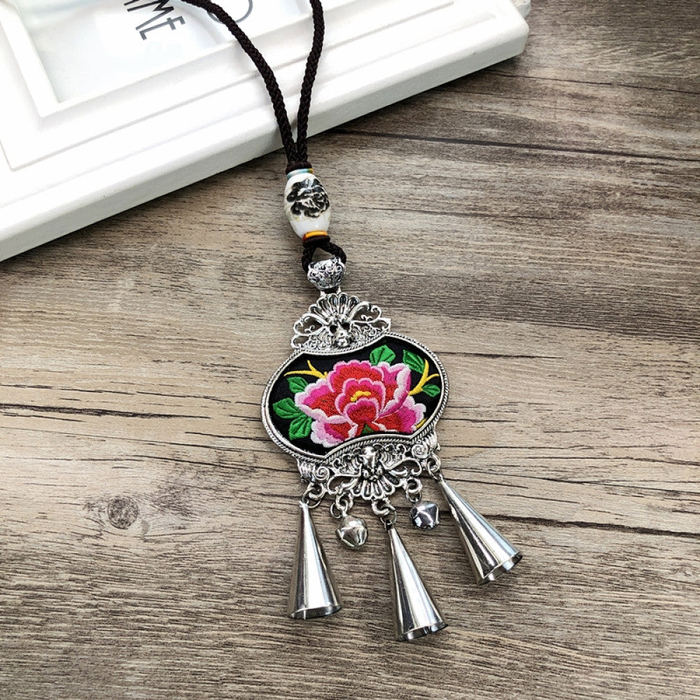 Vintage bohemian ethnic necklace with embroidered floral pendant, silver bells, and ceramic accent, displayed on a wooden surface with flowers, ideal for boho fashion enthusiasts 02