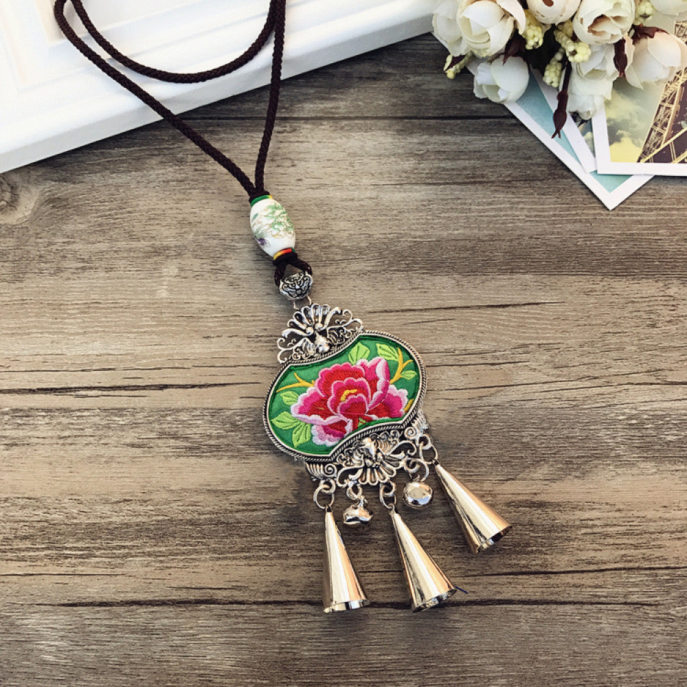 Vintage bohemian ethnic necklace with embroidered floral pendant, silver bells, and ceramic accent, displayed on a wooden surface with flowers, ideal for boho fashion enthusiasts 03