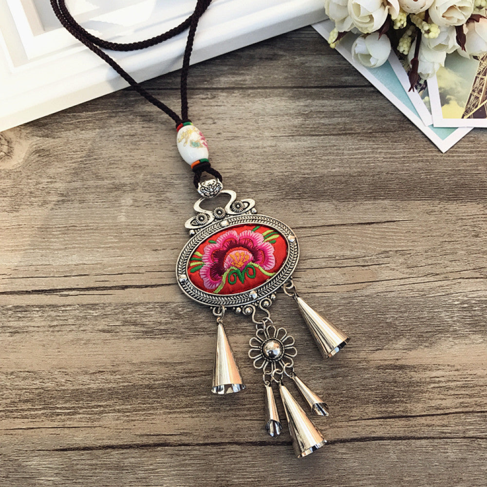Vintage bohemian ethnic necklace with embroidered floral pendant, silver bells, and ceramic accent, displayed on a wooden surface with flowers, ideal for boho fashion enthusiasts 05