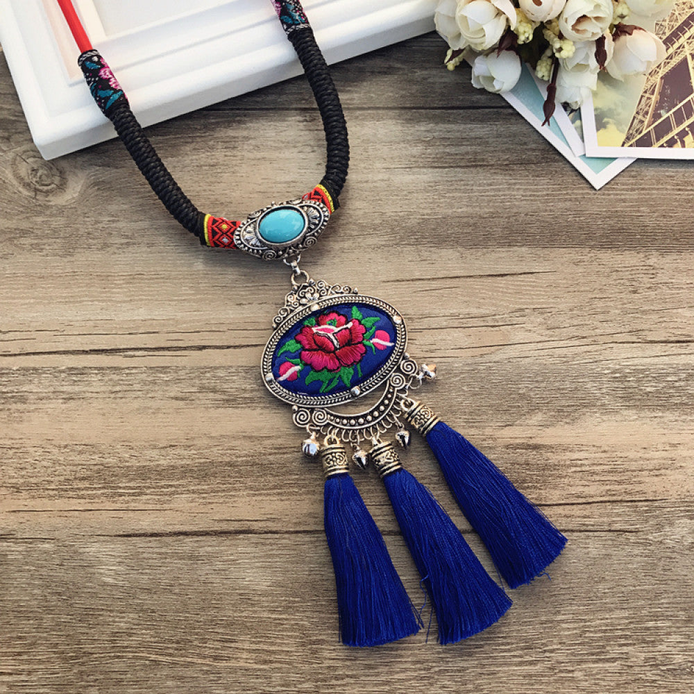 Vintage bohemian ethnic necklace with embroidered floral pendant, silver bells, and ceramic accent, displayed on a wooden surface with flowers, ideal for boho fashion enthusiasts 06