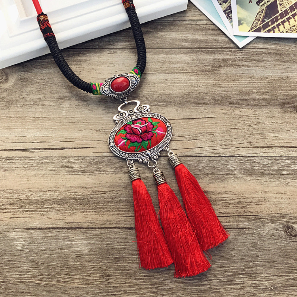 Vintage bohemian ethnic necklace with embroidered floral pendant, silver bells, and ceramic accent, displayed on a wooden surface with flowers, ideal for boho fashion enthusiasts 07