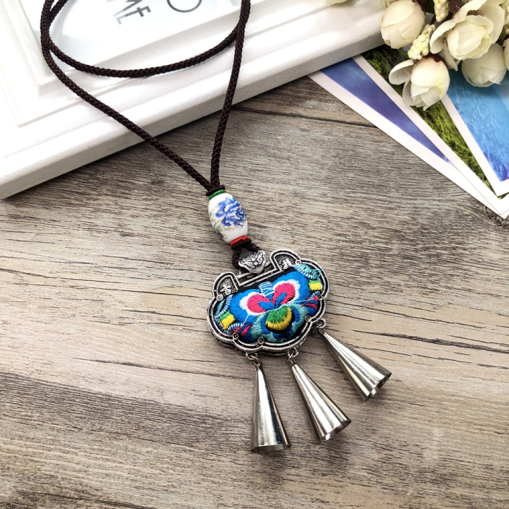 Vintage bohemian ethnic necklace with embroidered floral pendant, silver bells, and ceramic accent, displayed on a wooden surface with flowers, ideal for boho fashion enthusiasts 09