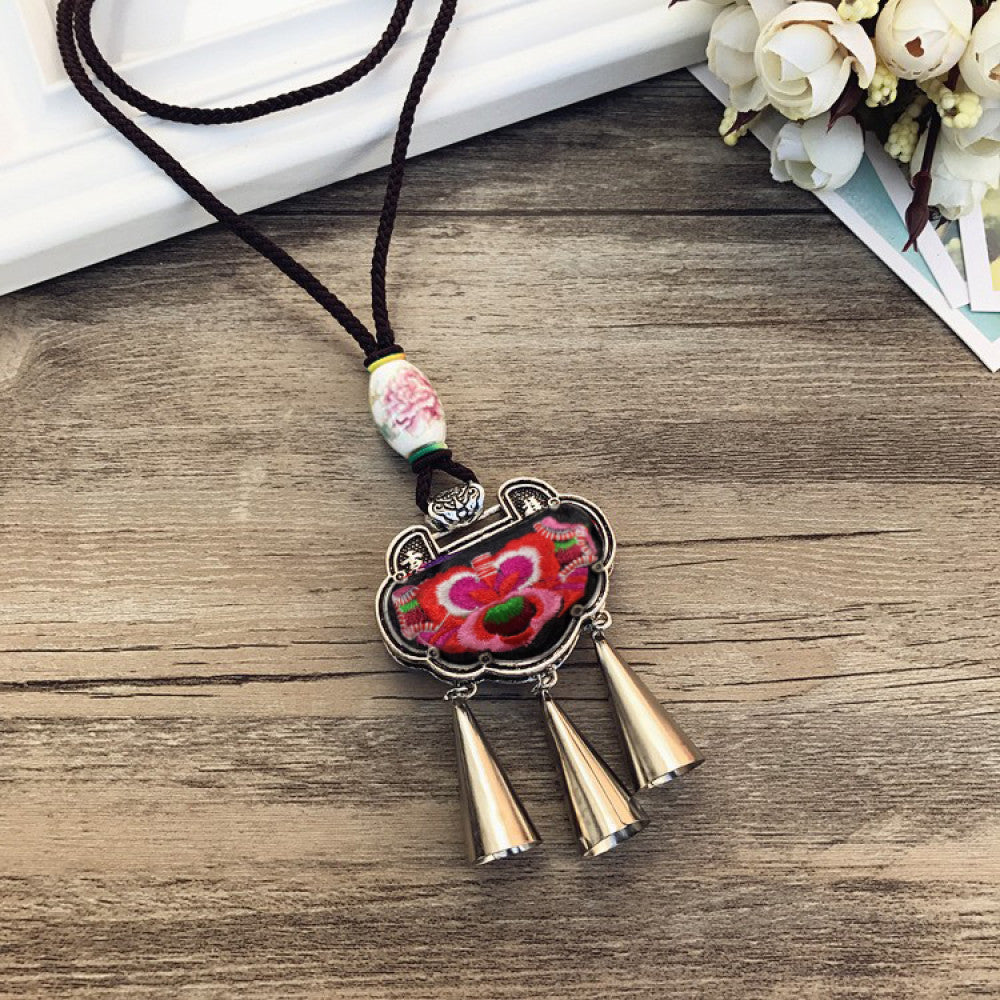Vintage bohemian ethnic necklace with embroidered floral pendant, silver bells, and ceramic accent, displayed on a wooden surface with flowers, ideal for boho fashion enthusiasts 10