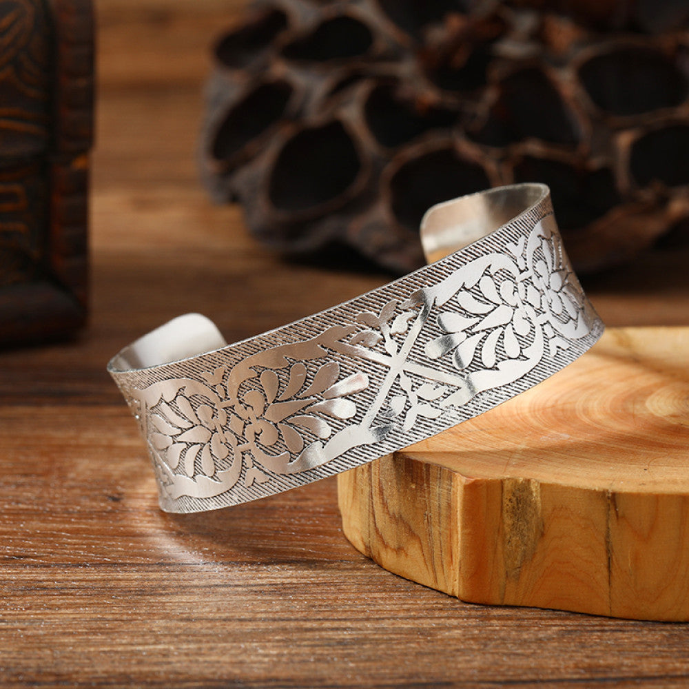 Vintage bohemian ethnic silver-tone cuff bracelet with intricate engraved floral designs, displayed on a wooden surface with rustic decor, ideal for boho and ethnic fashion enthusiasts