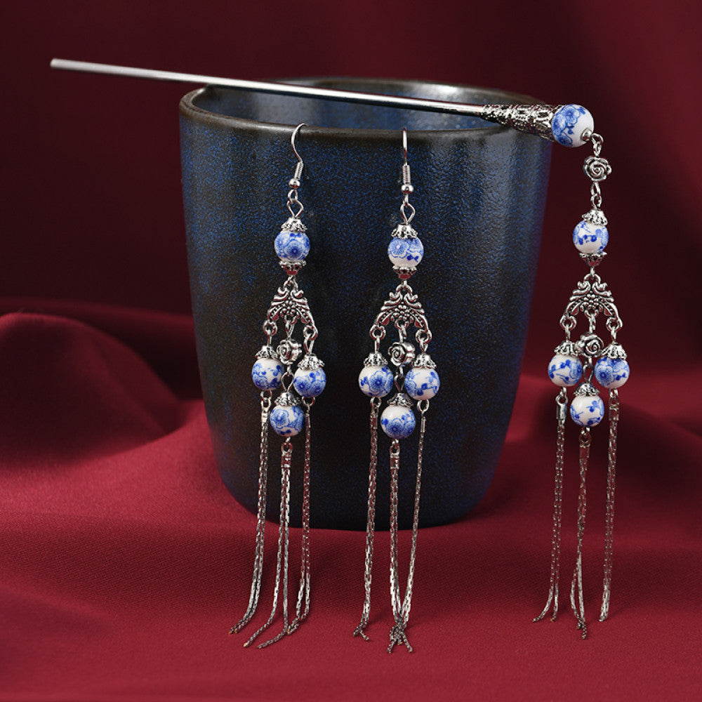 Boho vintage ethnic 3-piece set (1 hair stick + 2 dangle earrings) featuring silver-tone decor, blue-and-white porcelain-style beads, and layered chain tassel fringe, displayed beside a dark blue vessel on a rich red fabric background, ideal for bohemian and traditional fashion styling in the US