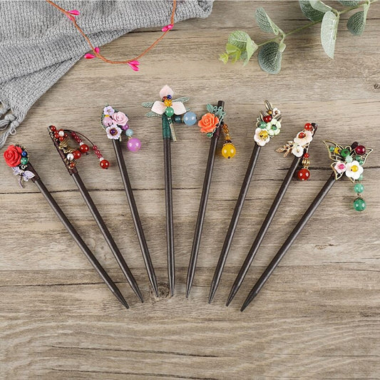 Set of 7 boho vintage ethnic dark wooden-look hair sticks, each featuring unique colorful flower charms & multi-color bead accents (pink, blue, orange, white, etc.), displayed on a rustic wooden surface (paired with gray fabric & green foliage decor), ideal for bohemian and traditional fashion enthusiasts in the US