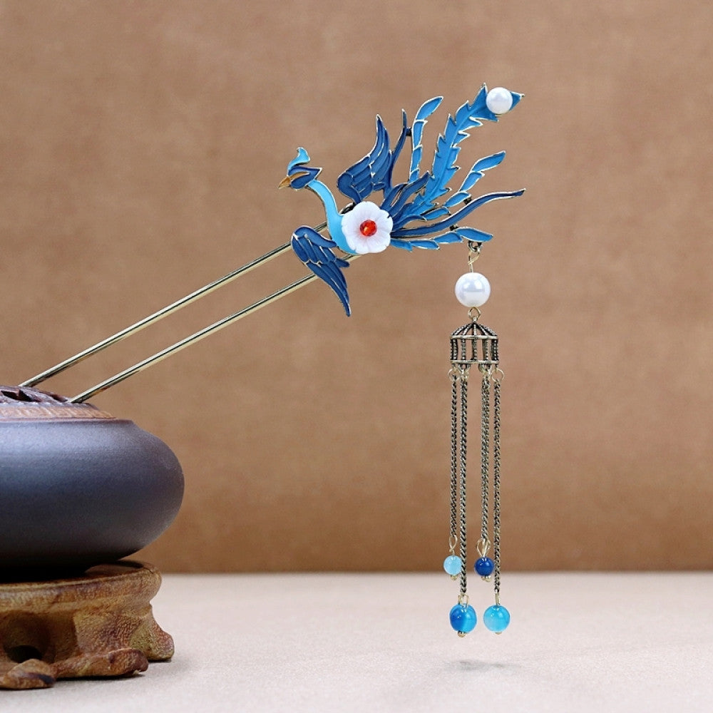 Boho vintage ethnic gold-tone double hair stick featuring a blue phoenix charm (with white flower & red center), pearl accent, birdcage pendant, and blue/white beaded tassels, displayed alongside a ceramic vessel on a warm brown background, ideal for bohemian and traditional fashion enthusiasts in the US