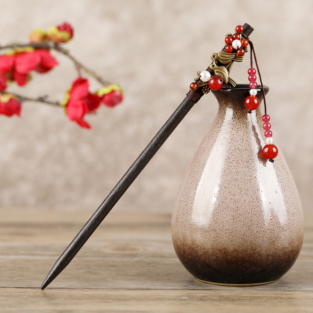 Boho vintage ethnic hair stick featuring a dark wooden-look handle, topped with light green flower, red bead, and green leaf decorative accents, displayed beside a red carved wooden decor 01