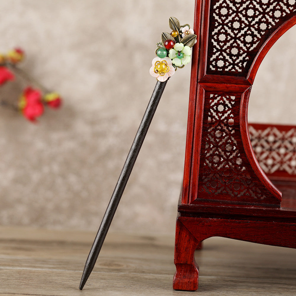 Boho vintage ethnic hair stick featuring a dark wooden-look handle, topped with light green flower, red bead, and green leaf decorative accents, displayed beside a red carved wooden decor  02