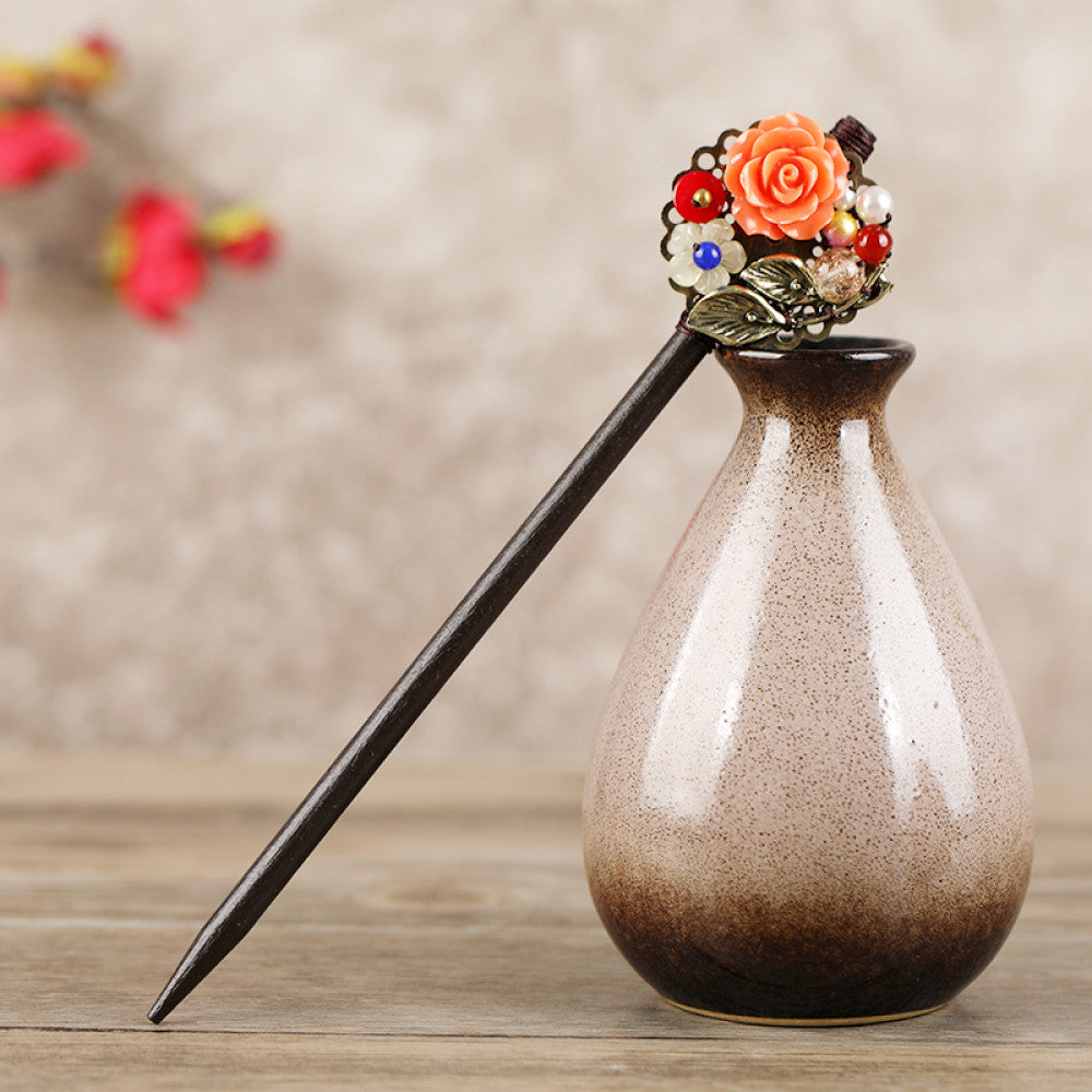 Boho vintage ethnic hair stick featuring a dark wooden-look handle, topped with light green flower, red bead, and green leaf decorative accents, displayed beside a red carved wooden decor  04