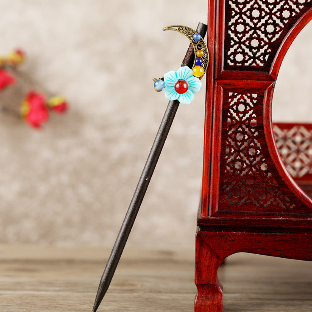 Boho vintage ethnic hair stick featuring a dark wooden-look handle, topped with light green flower, red bead, and green leaf decorative accents, displayed beside a red carved wooden decor 