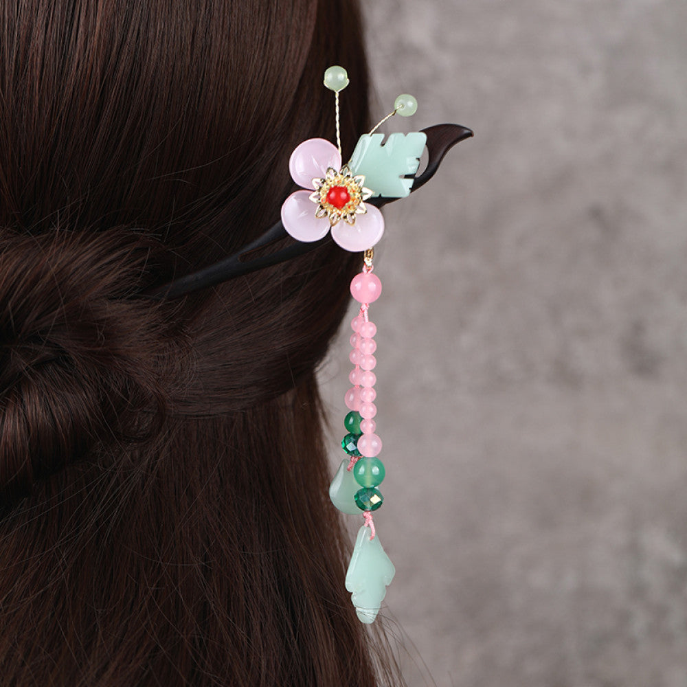 Boho vintage ethnic hair stick ornament (clipped in dark styled hair) featuring a pink flower (red center), green leaf accents, pink/green bead tassel, and light green leaf pendant, displayed against a muted gray background, ideal for bohemian traditional fashion styling in the US