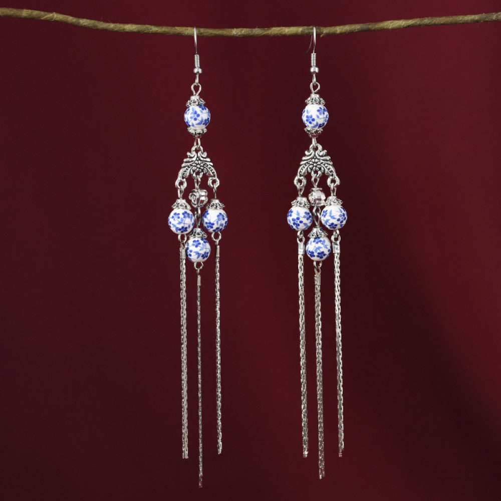Pair of boho vintage ethnic silver-tone dangle earrings, featuring blue-and-white porcelain-style beads, intricate decorative details, and layered chain tassel fringe, hung on a twig against a rich red fabric background