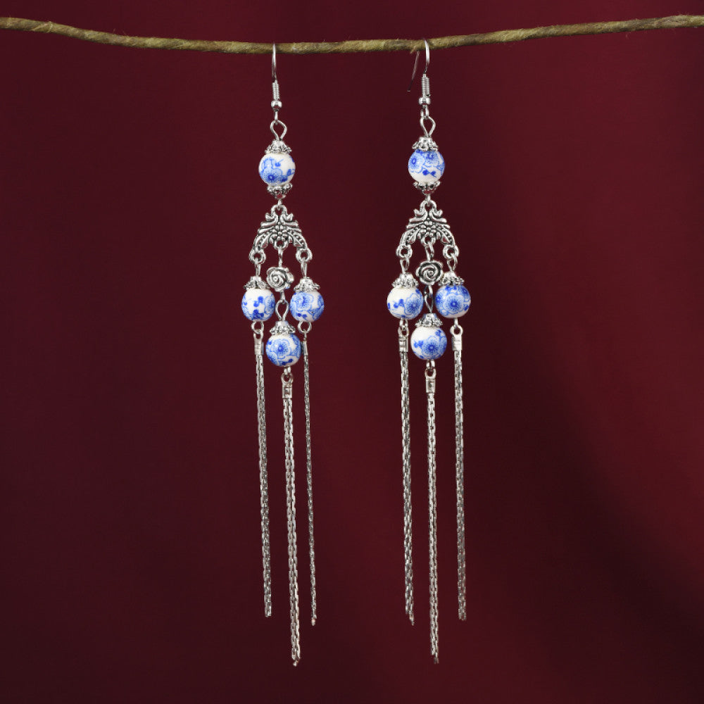 Pair of boho vintage ethnic silver-tone dangle earrings, featuring blue-and-white porcelain-style beads, intricate decorative details, and layered chain tassel fringe, hung on a twig against a rich red fabric background, ideal for bohemian and traditional fashion styling in the US