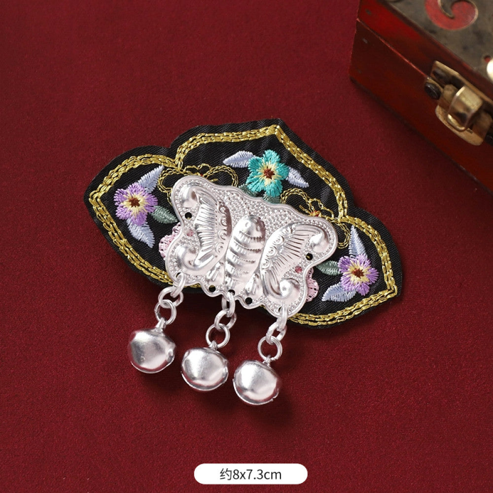 Boho vintage ethnic silver-tone decorative brooch featuring an intricate butterfly charm, pink floral embroidered accent (with blue details), and bell tassel pendants, displayed on a red fabric background  01