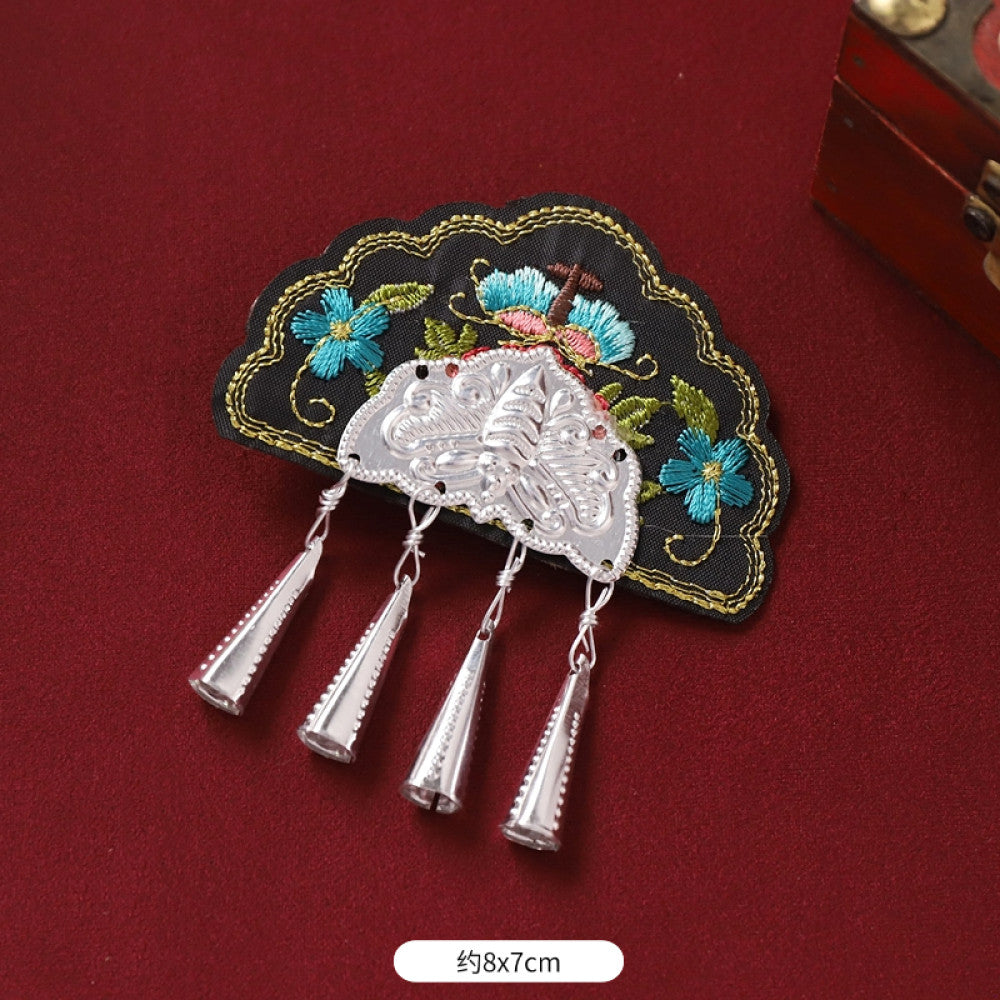Boho vintage ethnic silver-tone decorative brooch featuring an intricate butterfly charm, pink floral embroidered accent (with blue details), and bell tassel pendants, displayed on a red fabric background 05