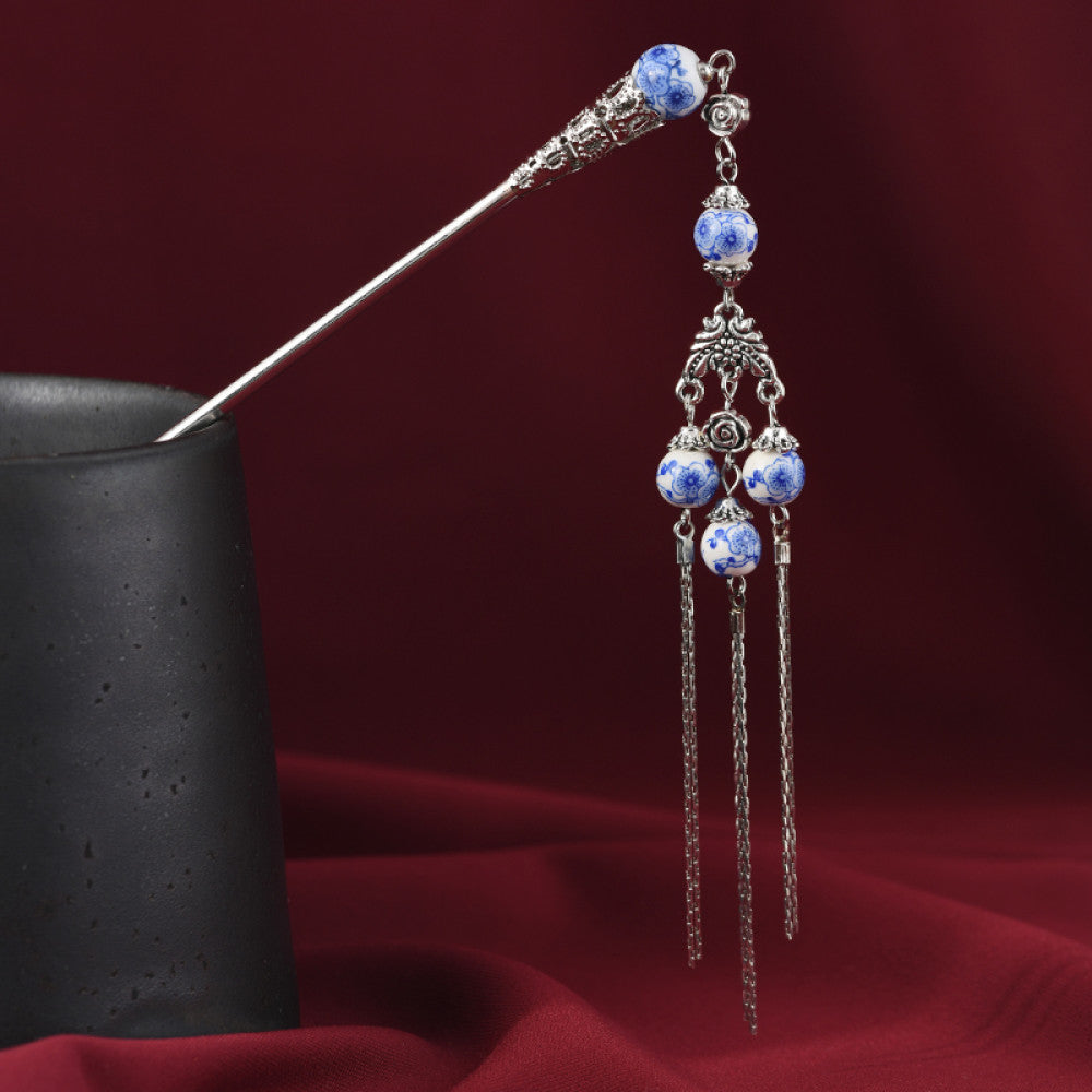 Boho vintage ethnic silver-tone hair stick featuring blue-and-white porcelain-style beads, intricate decorative details, and layered chain tassel fringe, displayed with a black vessel on a rich red fabric background, ideal for bohemian and traditional fashion enthusiasts in the US