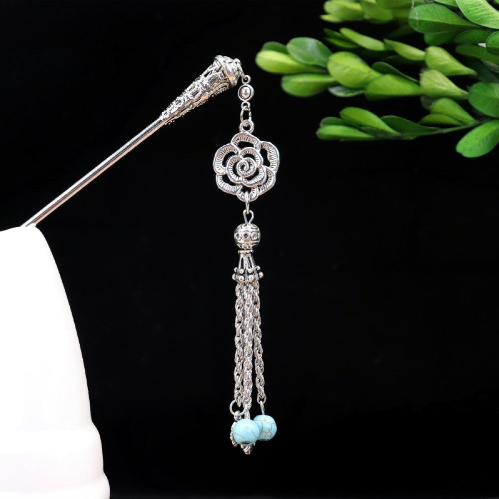 Boho vintage ethnic silver-tone hair stick featuring turquoise beads, an intricate round decorative element, and layered chain tassel fringe, displayed beside a white vessel green foliage