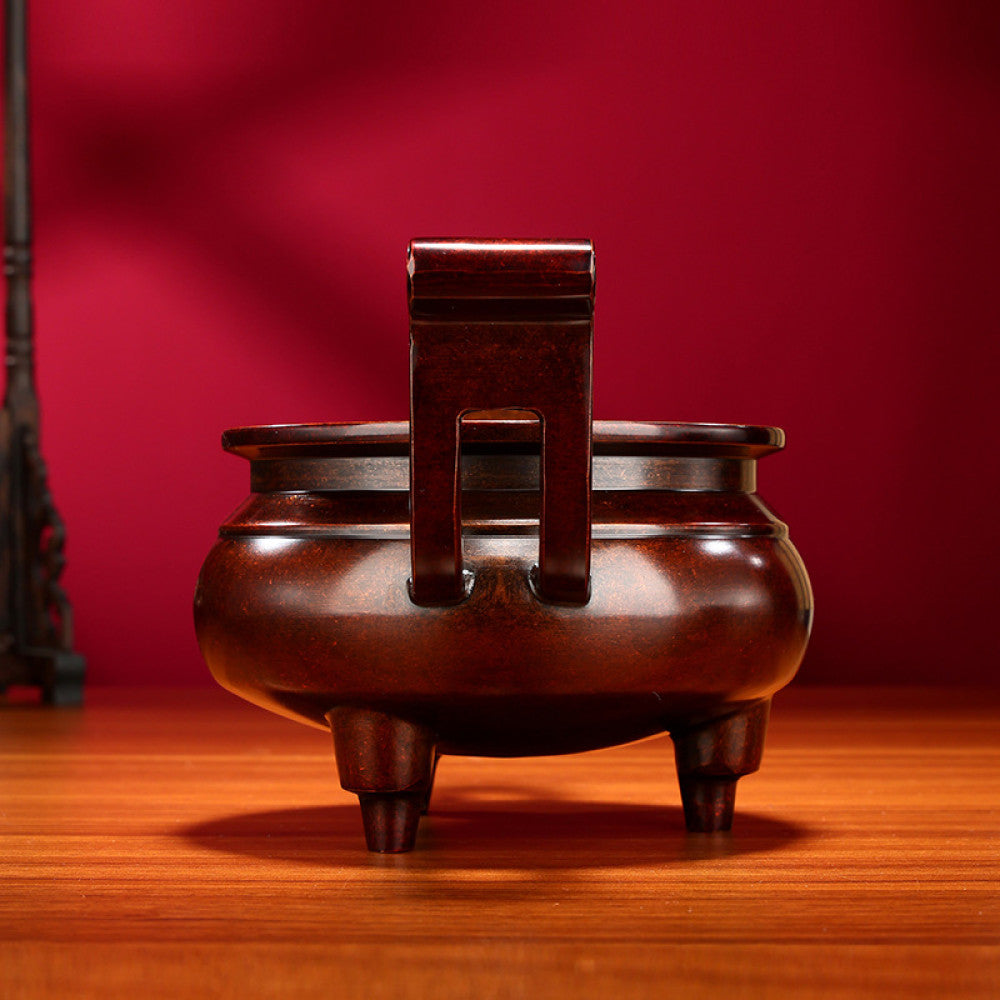 Intricate bronze Chinese incense burner with traditional curved handles and three feet, a Feng Shui symbol of wealth and harmony, displayed as elegant home decor on a wooden surface against a red background