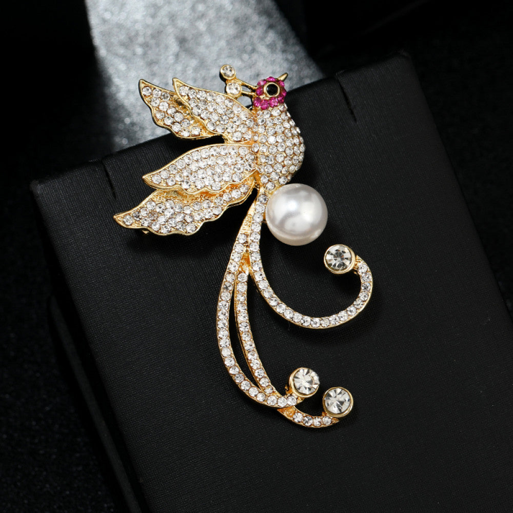 This elegant brooch features a sparkling crystal-encrusted bird in a gold-tone setting, accented by a lustrous pearl and delicate pink detail at its head. A sophisticated statement piece for any outfit