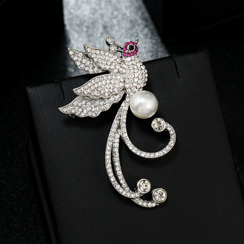 This elegant brooch features a sparkling crystal-encrusted bird in a gold-tone setting, accented by a lustrous pearl and delicate pink detail at its head. A sophisticated statement piece for any outfit 01