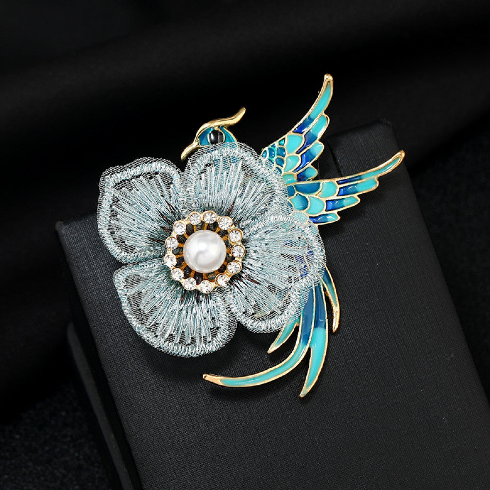 This exquisite brooch features a graceful enamel bird in vibrant blue hues, perched beside a delicate lace flower. The flower’s center is adorned with a lustrous pearl, surrounded by a halo of sparkling crystals, creating a stunning contrast of textures and colors. Perfect for adding a touch of whimsical elegance to blazers, scarves, or special occasion outfits, this piece blends intricate craftsmanship with timeless, eye-catching charm