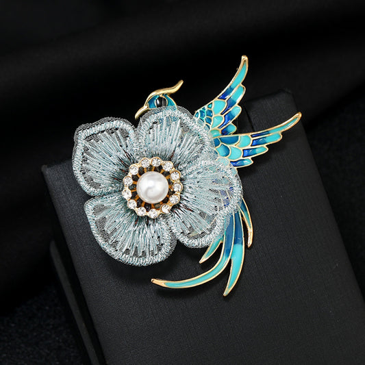 This exquisite brooch features a graceful enamel bird in vibrant blue hues, perched beside a delicate lace flower. The flower’s center is adorned with a lustrous pearl, surrounded by a halo of sparkling crystals, creating a stunning contrast of textures and colors. Perfect for adding a touch of whimsical elegance to blazers, scarves, or special occasion outfits, this piece blends intricate craftsmanship with timeless, eye-catching charm