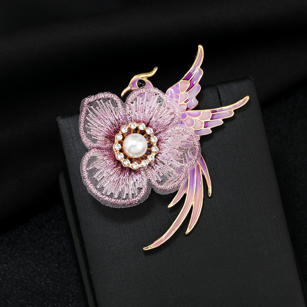 This exquisite brooch features a graceful enamel bird in vibrant blue hues, perched beside a delicate lace flower. The flower’s center is adorned with a lustrous pearl, surrounded by a halo of sparkling crystals, creating a stunning contrast of textures and colors. Perfect for adding a touch of whimsical elegance to blazers, scarves, or special occasion outfits, this piece blends intricate craftsmanship with timeless, eye-catching charm 01