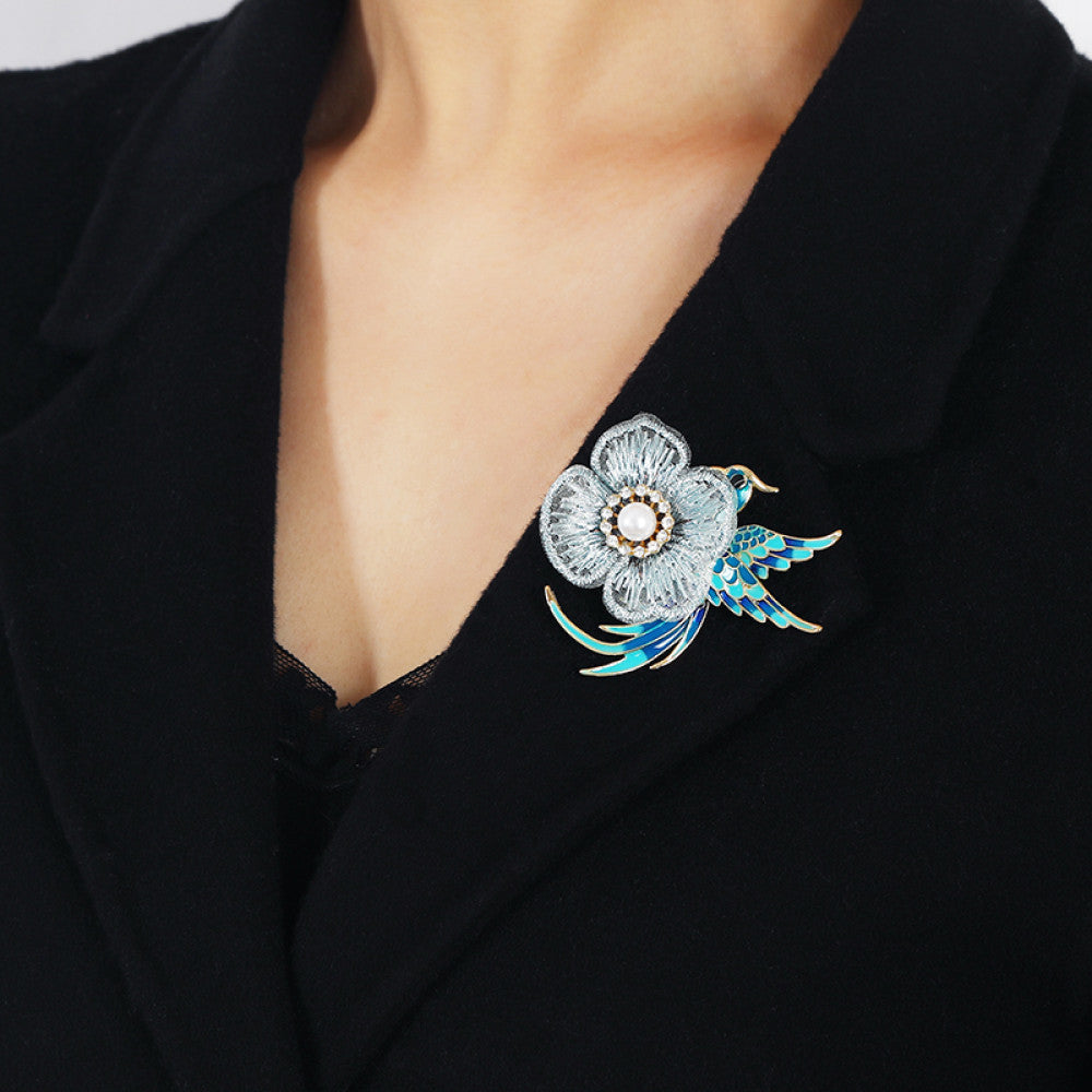 This exquisite brooch features a graceful enamel bird in vibrant blue hues, perched beside a delicate lace flower. The flower’s center is adorned with a lustrous pearl, surrounded by a halo of sparkling crystals, creating a stunning contrast of textures and colors. Perfect for adding a touch of whimsical elegance to blazers, scarves, or special occasion outfits, this piece blends intricate craftsmanship with timeless, eye-catching charm 02
